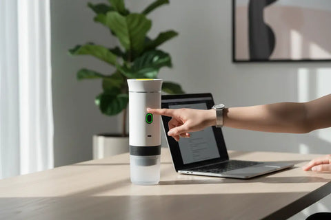 Cafetera portátil 3 en 1 sobre escritorio de oficina junto a portátil, ideal para preparar café en el trabajo.