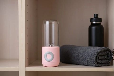 Exprimidor eléctrico portátil rosa con vaso de cristal transparente, junto a toalla enrollada gris y botella negra en estantería de madera